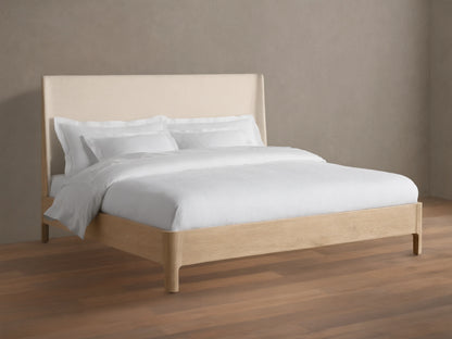 Front view of the Calista Bed, featuring a light oak wood frame and a cream upholstered headboard, dressed with white bedding.