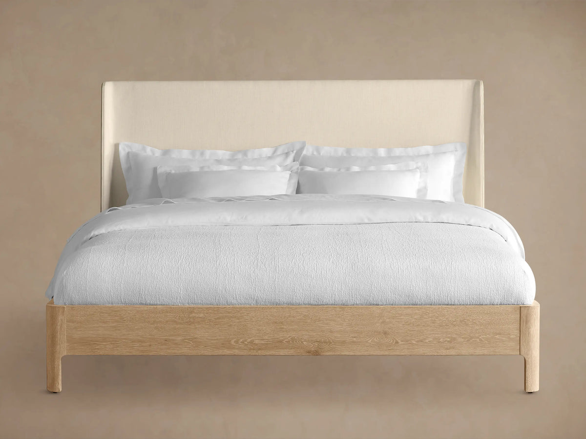Front view of the Calista Bed, featuring a light oak wood frame and a cream upholstered headboard, dressed with white bedding.