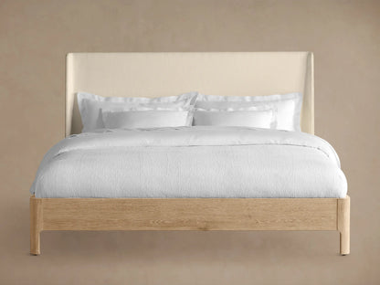 Front view of the Calista Bed, featuring a light oak wood frame and a cream upholstered headboard, dressed with white bedding.
