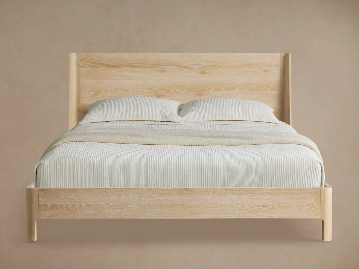 Calista Wood Queen Bed in sand finish with a tall, angled headboard, and neatly made in neutral bedding, on wood flooring.