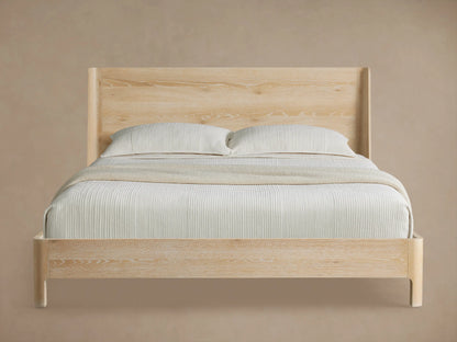 Calista Wood Queen Bed in sand finish with a tall, angled headboard, and neatly made in neutral bedding, on wood flooring.