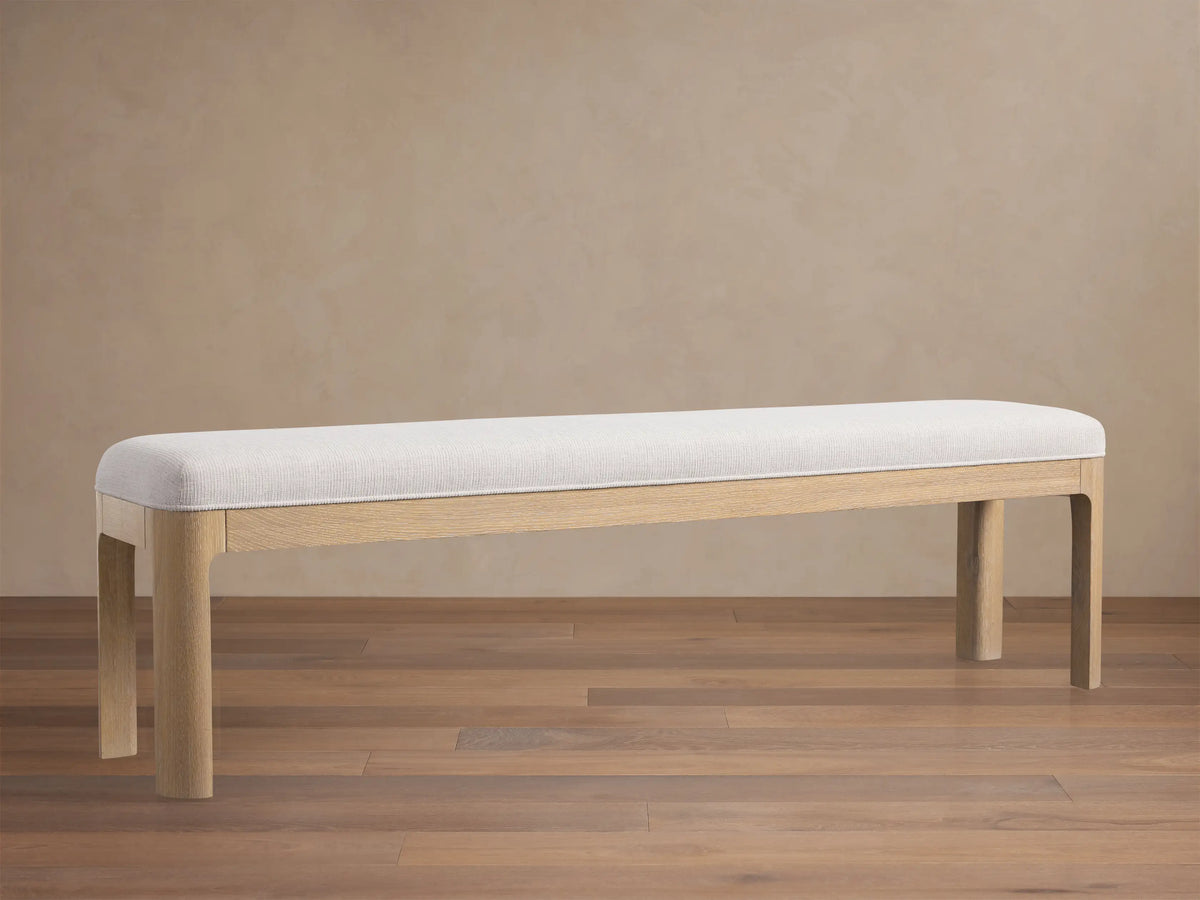 Calista Bench: a light oak bench with a light, subtly textured upholstered top, centered on a wood floor against a beige wall.