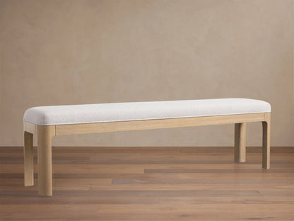 Calista Bench: a light oak bench with a light, subtly textured upholstered top, centered on a wood floor against a beige wall.