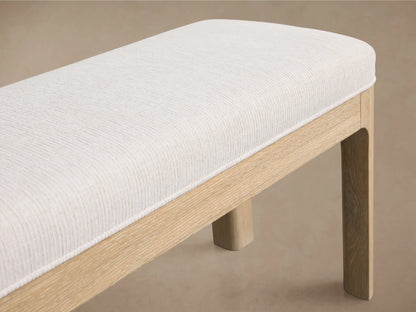 Calista Bench. Light oak wood frame, slender legs, and long, textured cream upholstered seat. Against a warm beige wall.