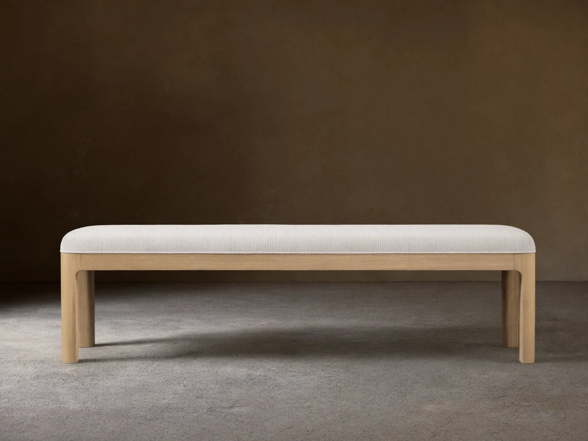 Calista Bench: a light oak bench with a light, subtly textured upholstered top, centered on a wood floor against a beige wall.