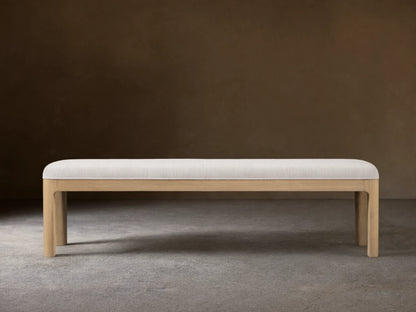 Calista Bench: a light oak bench with a light, subtly textured upholstered top, centered on a wood floor against a beige wall.
