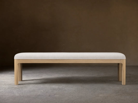 Calista Bench: a light oak bench with a light, subtly textured upholstered top, centered on a wood floor against a beige wall.