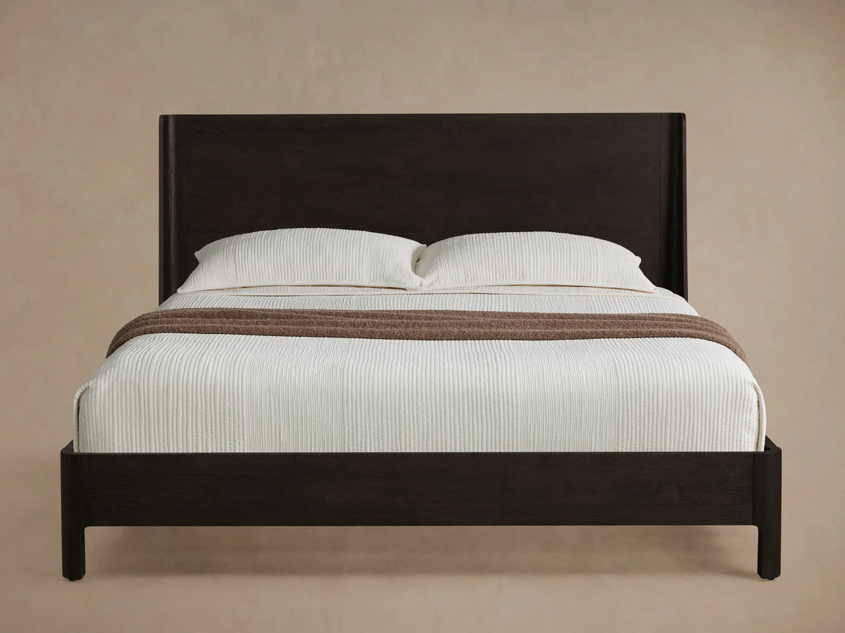 Calista Wood Bed, dark brown oak frame with simple headboard, white bedding, and brown throw blanket on a wood floor.