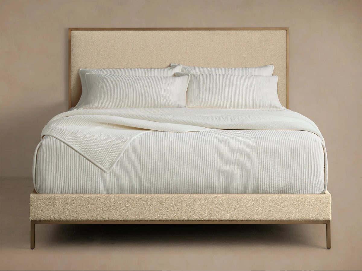 Beige Chamberlain Bed with upholstered headboard and frame, gold trim. White striped quilt and pillows on top.