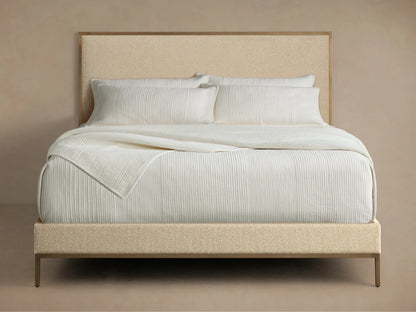 Beige Chamberlain Bed with upholstered headboard and frame, gold trim. White striped quilt and pillows on top.