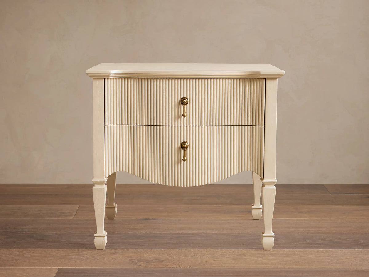 Cream Conrad Nightstand with reeded facade, 2 drawers, antique brass pulls, and curved base. Tapered legs on wood floor.