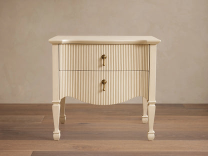 Cream Conrad Nightstand with reeded facade, 2 drawers, antique brass pulls, and curved base. Tapered legs on wood floor.