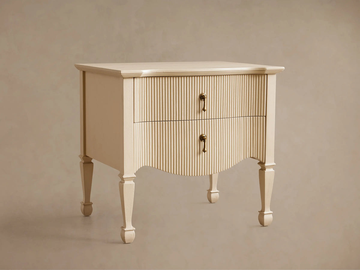 Cream Conrad Nightstand with reeded facade, 2 drawers, antique brass pulls, and curved base. Tapered legs on wood floor.