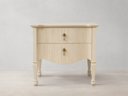 Cream Conrad Nightstand with reeded facade, 2 drawers, antique brass pulls, and curved base. Tapered legs on wood floor.
