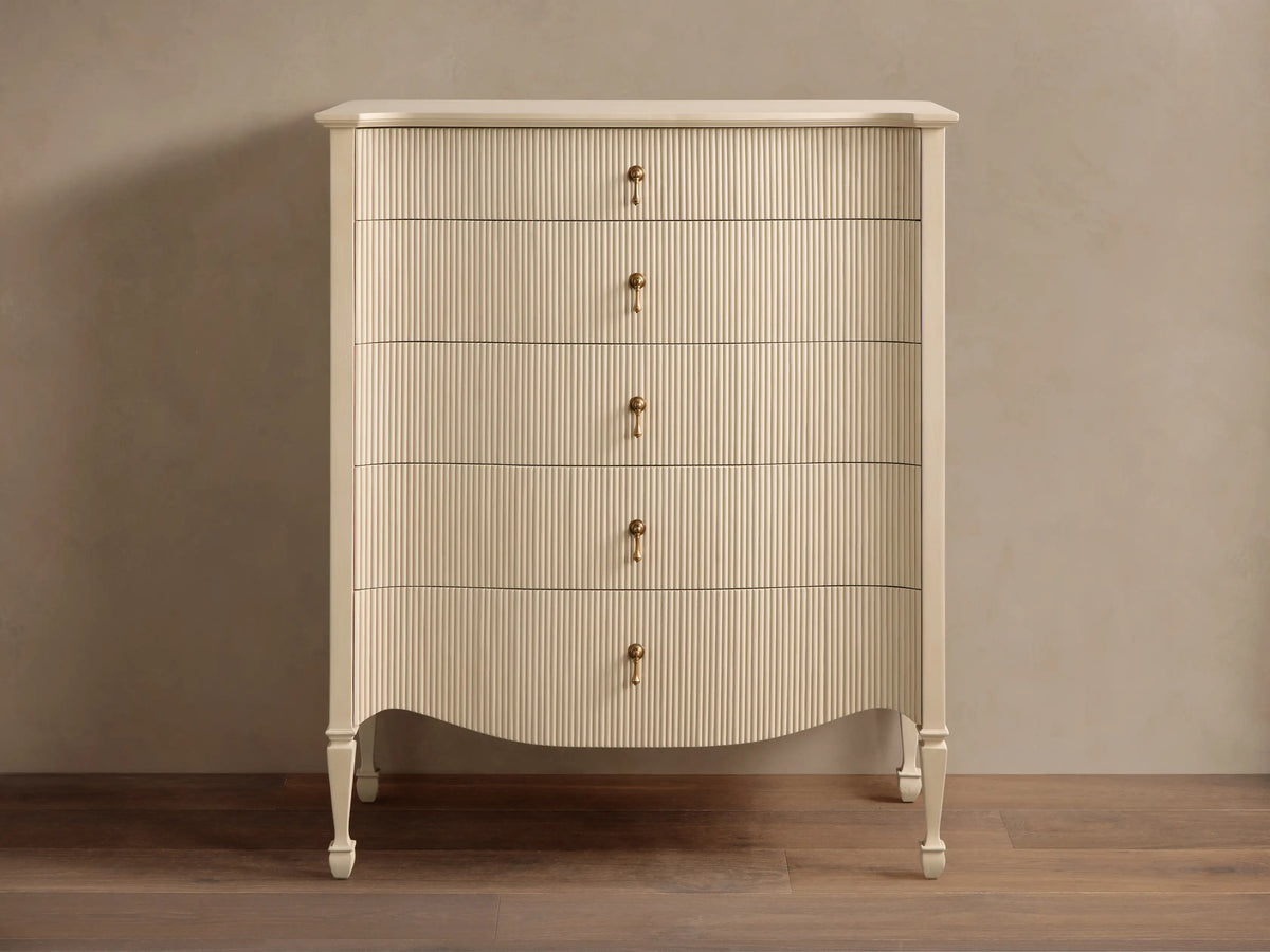 Conrad Five-Drawer Dresser, light cream, reeded fronts, 5 antique brass pendant pulls, curved base, on wooden floor.
