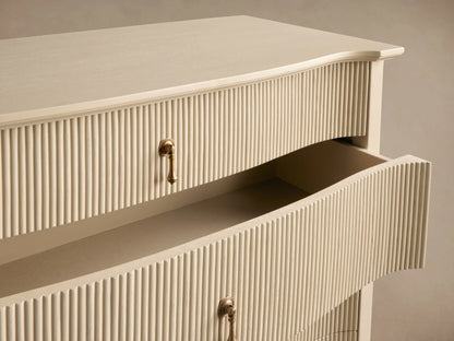Conrad Five-Drawer Dresser, light cream, reeded fronts, 5 antique brass pendant pulls, curved base, on wooden floor.