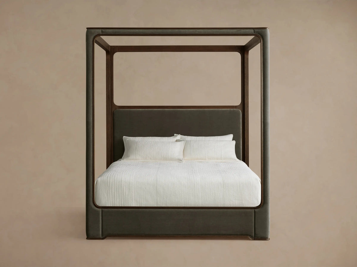 Full front view of the Conley Canopy Bed with a dark oak frame, upholstered dark brown headboard, and cream bedding. A small dog rests on the bed.