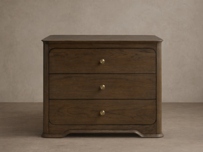 Dark oak Conley Nightstand with three drawers, champagne brass pulls, curved edges, and a sculpted base, shown front on.