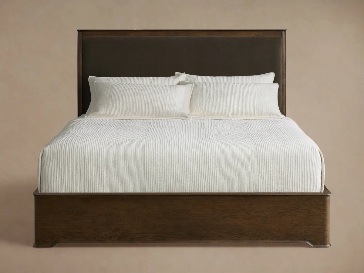 Conley Panel Bed with dark wood frame & upholstered headboard, dressed in crisp white bedding, against a beige wall & wood floor.