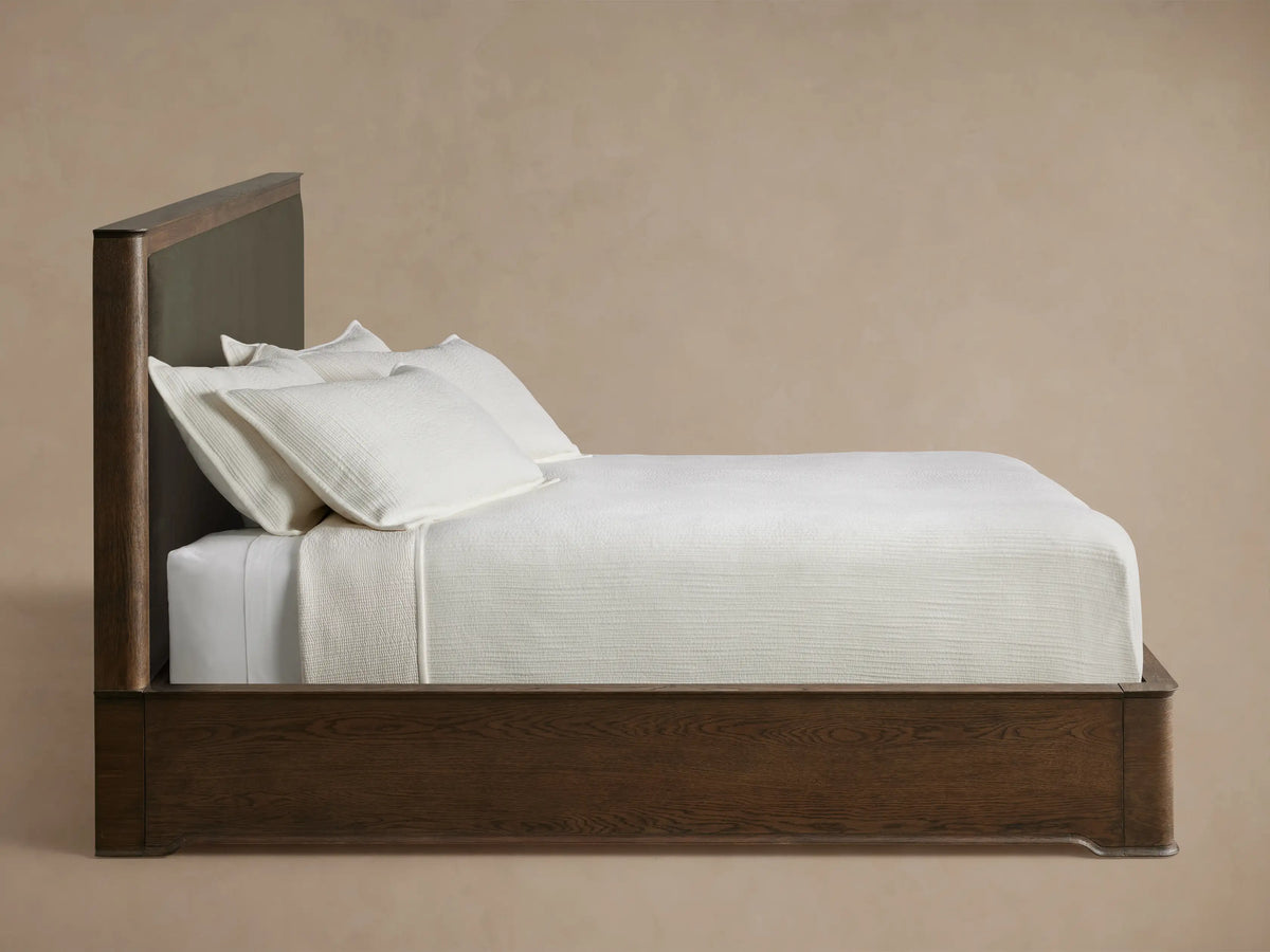 Dark oak wood frame with visible grain details curves around a dark olive upholstered panel of the Conley Panel Bed headboard.