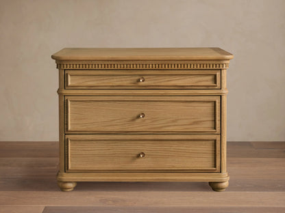 Carlisle Nightstand in light oak, front view. Three drawers with antique brass pulls, carved detailing below top, and round feet.