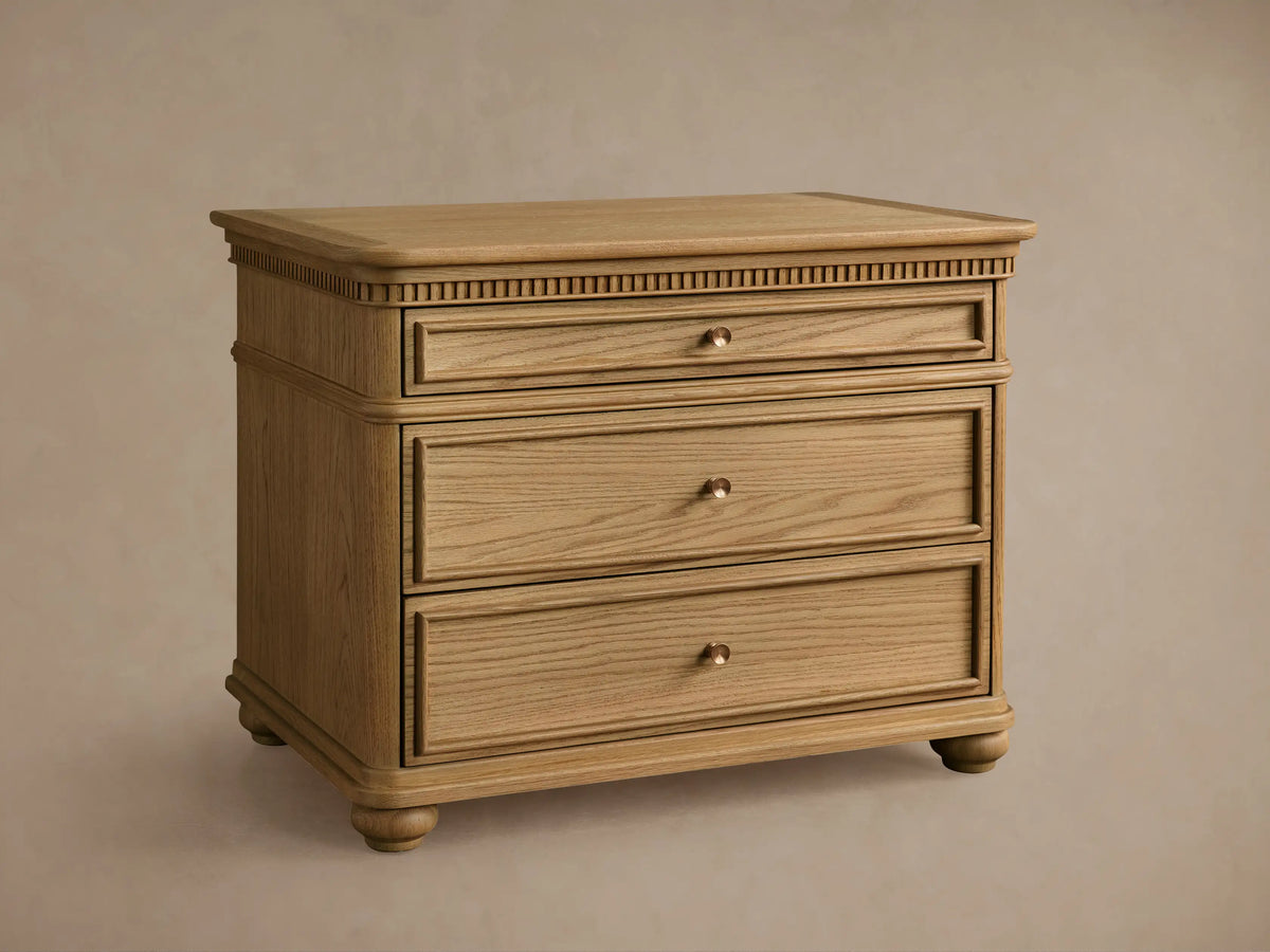 Carlisle Nightstand in light oak, front view. Three drawers with antique brass pulls, carved detailing below top, and round feet.