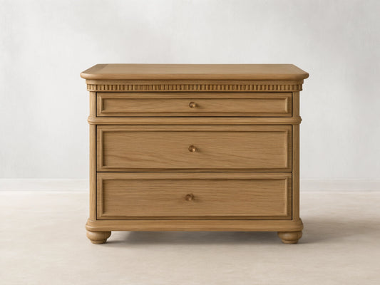 Carlisle Nightstand in light oak, front view. Three drawers with antique brass pulls, carved detailing below top, and round feet.