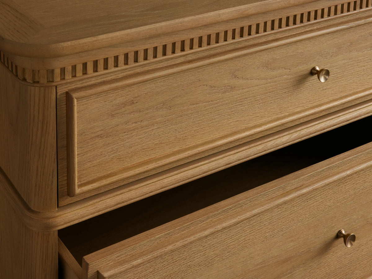 Tall Carlisle Five-Drawer Dresser, light oak wood, antique brass pulls, fluted trim above top drawer, against warm background.