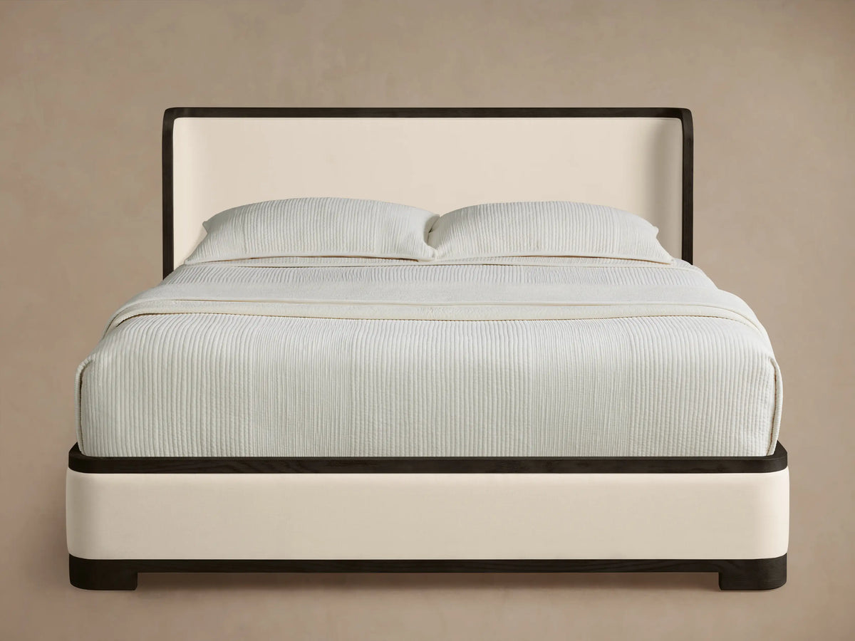 Darien Curved Shelter Bed, cream upholstered with dark frame, curved headboard & light ribbed bedding on a wood floor.