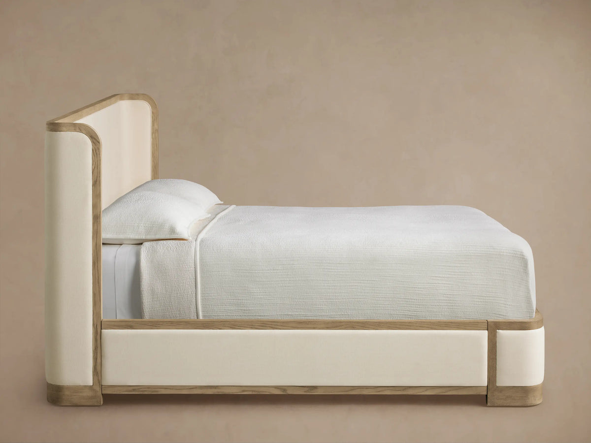 Close-up of Darien Curved Shelter Bed with a curved dark gray-brown oak wood frame with visible grain bordering a light beige upholstered panel.