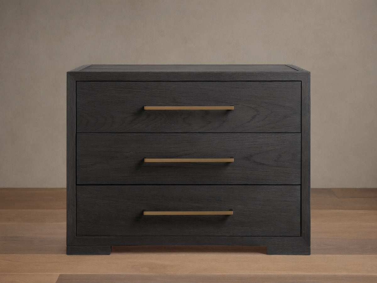 Darwyn Nightstand made of dark oak wood with three drawers featuring brass handles, sitting on a wooden floor.