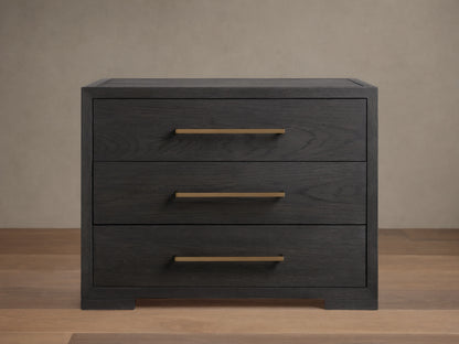 Darwyn Nightstand made of dark oak wood with three drawers featuring brass handles, sitting on a wooden floor.