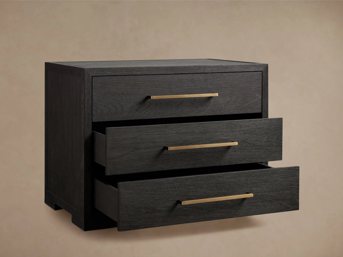 Darwyn Nightstand made of dark oak wood with three drawers featuring brass handles, sitting on a wooden floor.