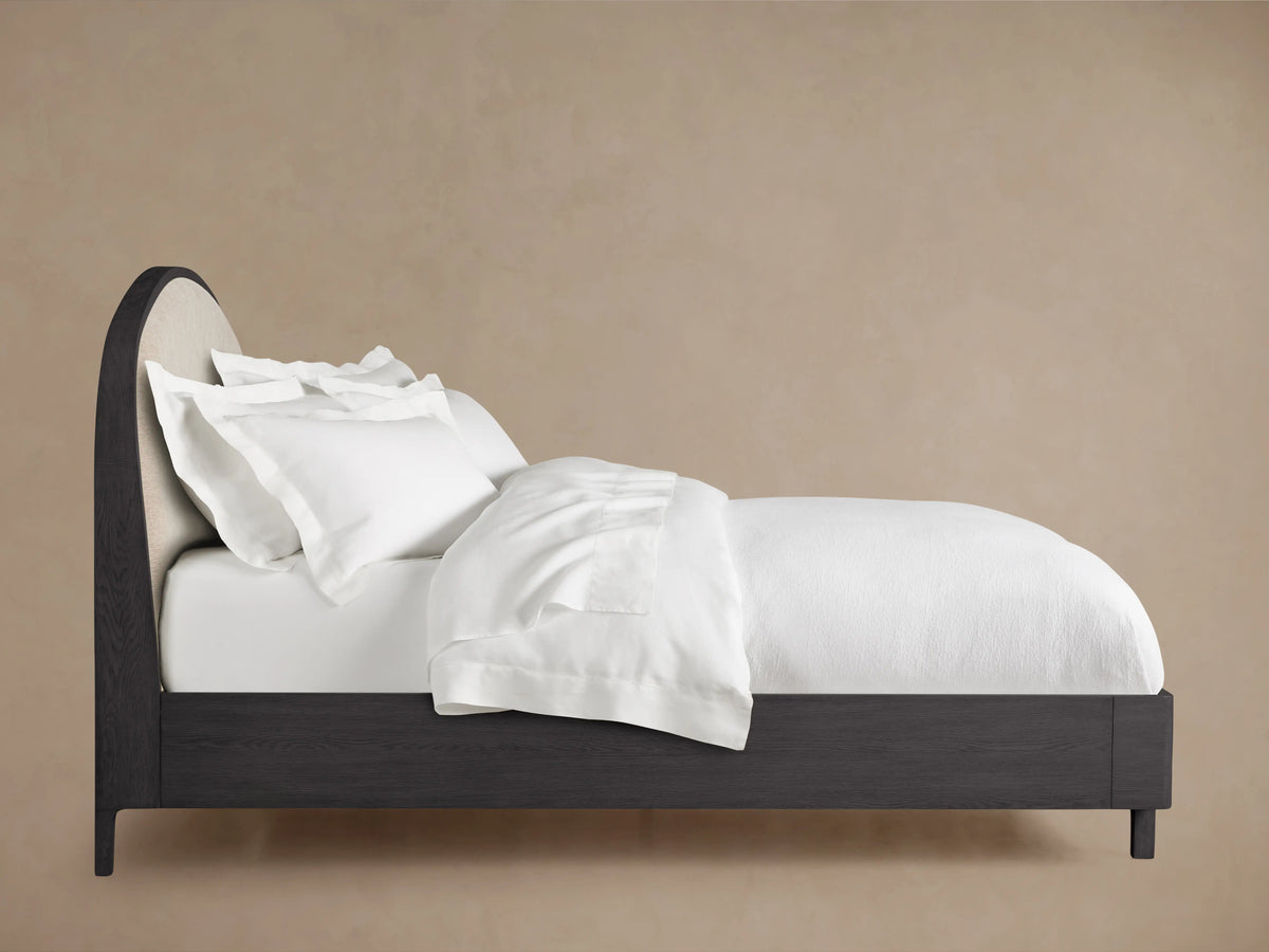 Danny Bed in ebony finish, featuring a curved upholstered headboard with white bedding and pillows, on a beige background.