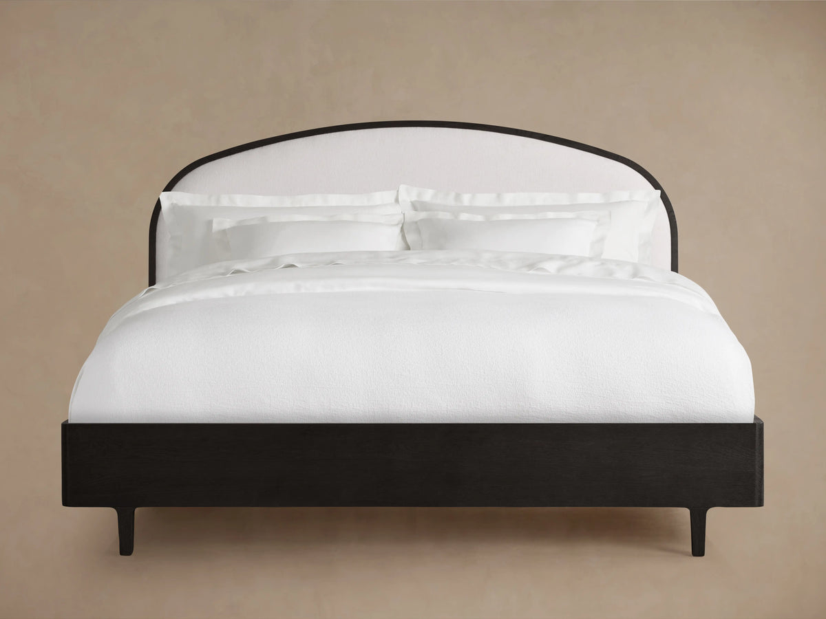 Danny Bed with an arched, upholstered white headboard and ebony-finished oak frame, set on a wooden floor in a neutral-toned room.