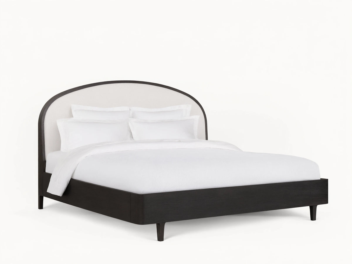 Danny Bed with an arched, upholstered white headboard and ebony-finished oak frame, set on a wooden floor in a neutral-toned room.