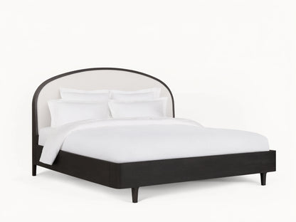 Danny Bed with an arched, upholstered white headboard and ebony-finished oak frame, set on a wooden floor in a neutral-toned room.
