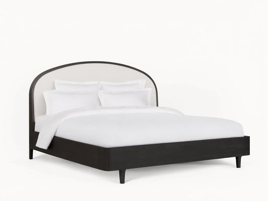 Danny Bed with an arched, upholstered white headboard and ebony-finished oak frame, set on a wooden floor in a neutral-toned room.