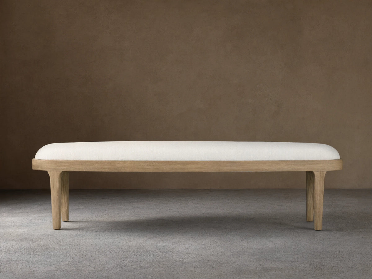 Danny Bench with a sleek, rectangular design, upholstered in light fabric, set on oak legs with a fawn finish, against a beige wall.