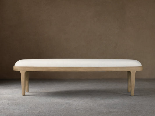 Danny Bench with a sleek, rectangular design, upholstered in light fabric, set on oak legs with a fawn finish, against a beige wall.