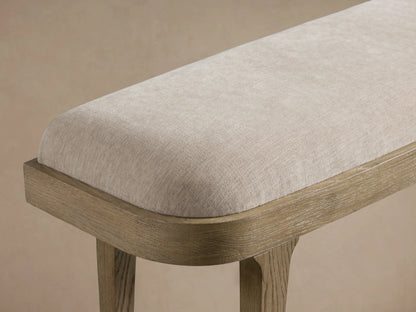 Danny Bench in fawn finish, upholstered in light beige fabric, with a wooden frame and four sturdy legs, on a wooden floor.