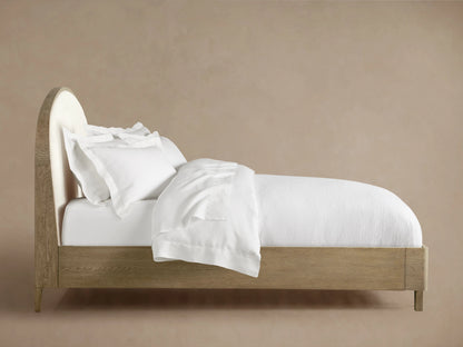Danny Bed with a rounded upholstered headboard in white, oak wood frame in a fawn finish, and neatly made white bedding.