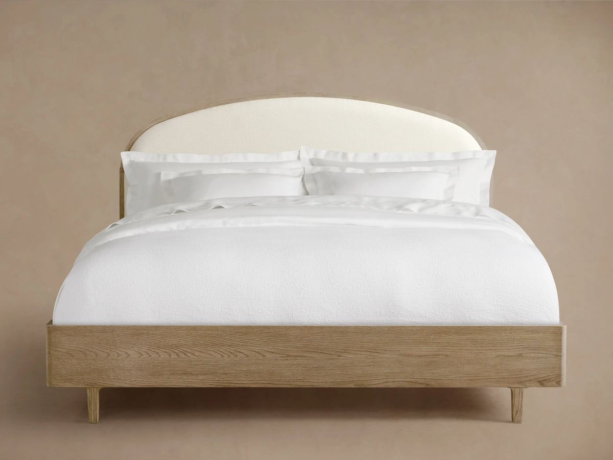 Danny Bed with a curved, upholstered headboard in white fabric. Oak wood frame with a fawn finish, neatly made with white bedding.