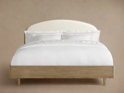 Danny Bed with a curved, upholstered headboard in white fabric. Oak wood frame with a fawn finish, neatly made with white bedding.