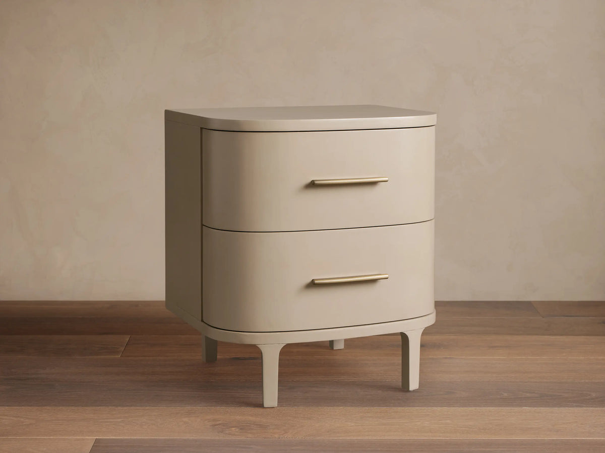 Danny Nightstand in fumo: light beige, rounded nightstand with two drawers, horizontal brass pulls, on short splayed legs.