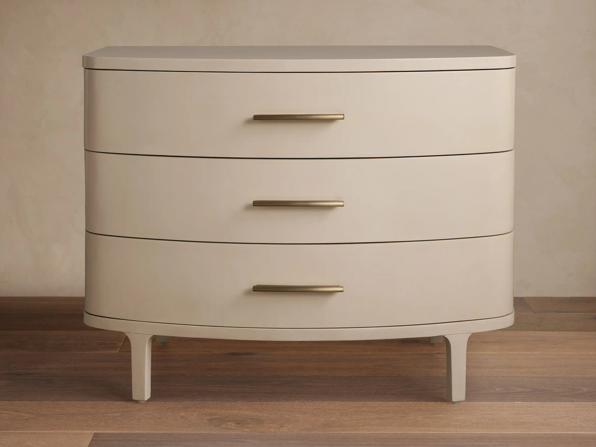 Danny Three-Drawer Chest in Fumo. Light grayish-beige finish, rounded front, 3 drawers with brass pulls, on wood floor.