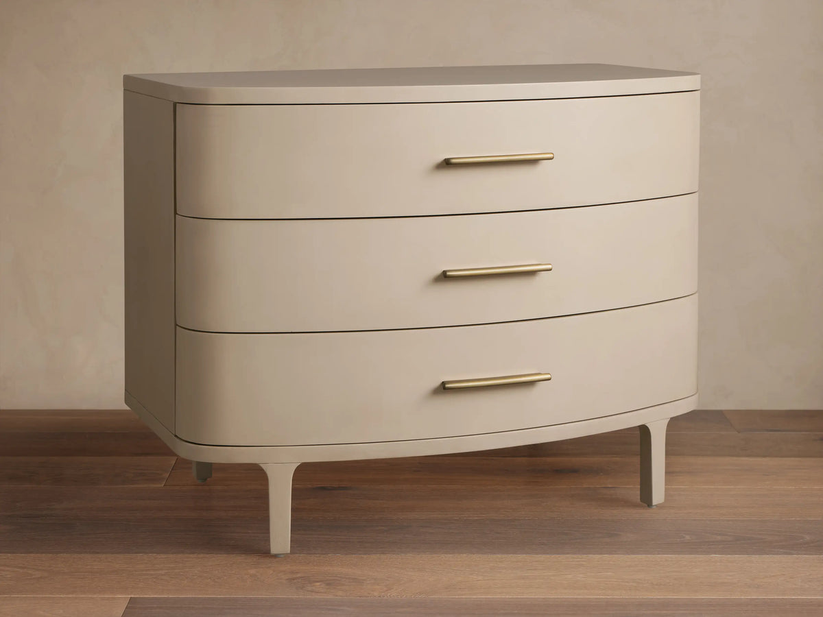Danny Three-Drawer Chest in Fumo. Light grayish-beige finish, rounded front, 3 drawers with brass pulls, on wood floor.