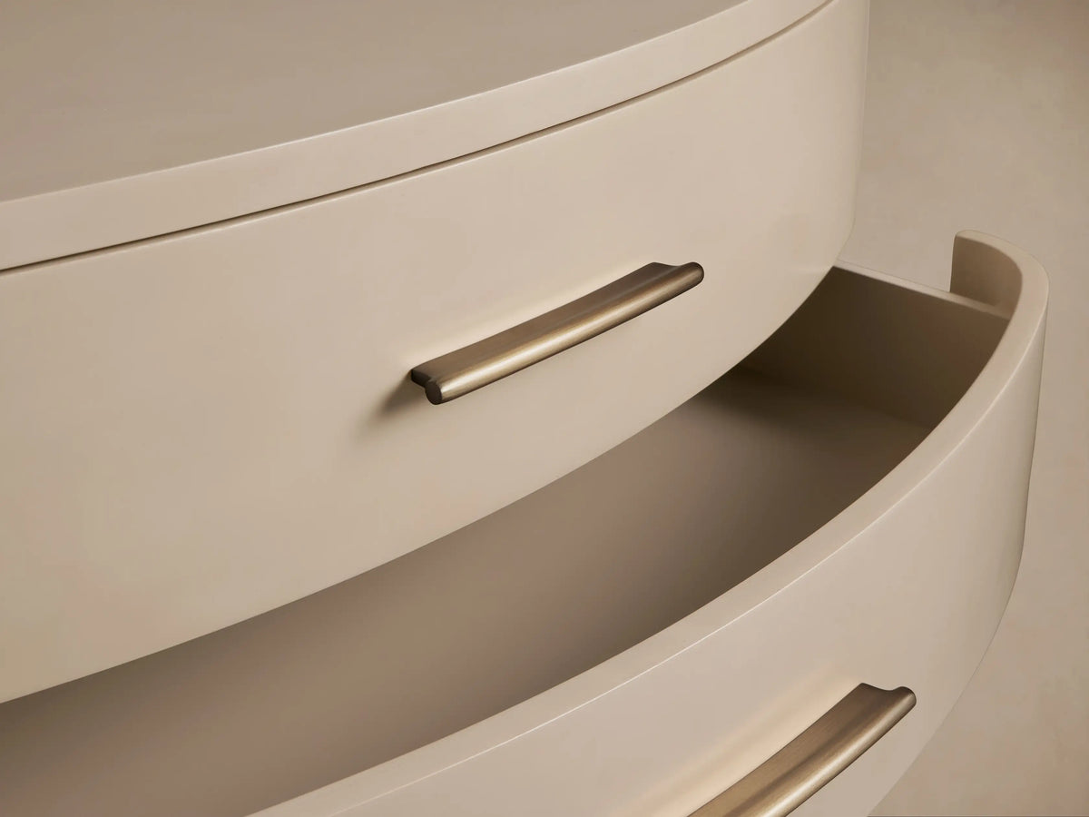Danny Fumo Three-Drawer Chest. Light greyish-beige chest with curved fronts, 3 brass pulls, short legs, on wooden floor.