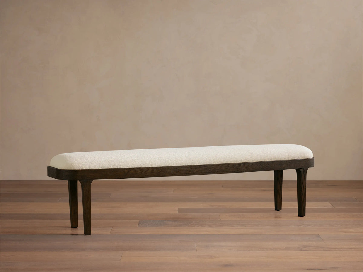 Danny Bench: Upholstered cream fabric top with rounded edges on a dark oak wooden frame & tapered legs. Full view in room.