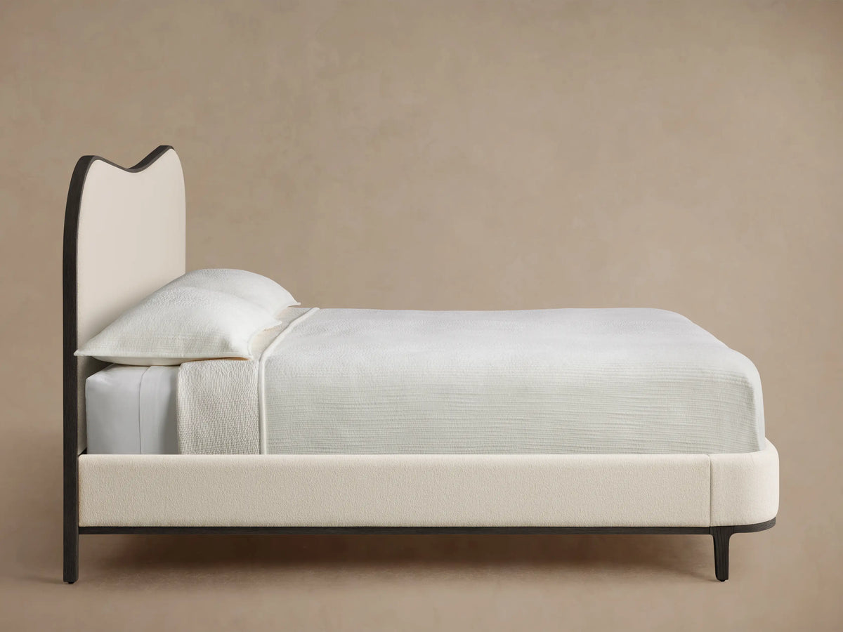 Close-up of a Danny Camelback Bed headboard's top right curve, featuring textured cream fabric and a dark, curved wooden frame.